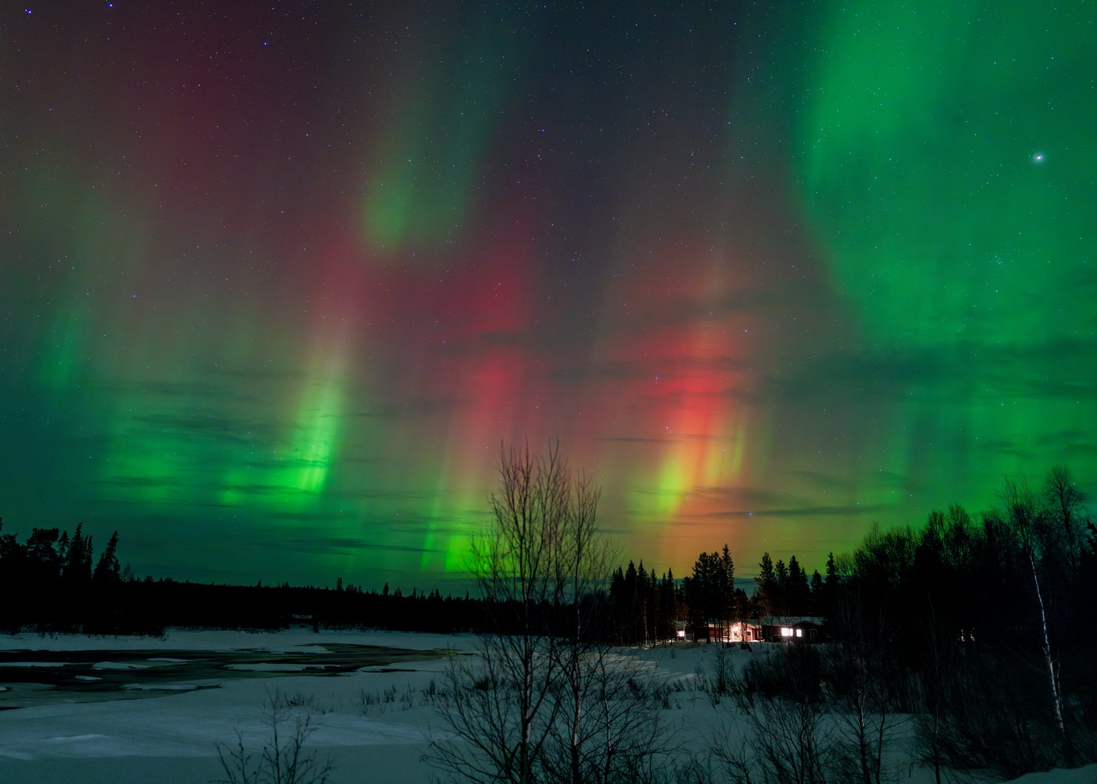Northern lights over a snowy field with a lit cabin