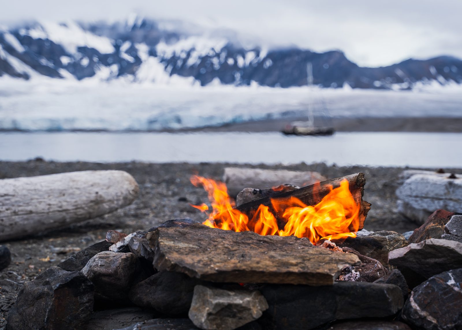 Campfire on a gravel beach, a sailboat in front of a glacier in the background